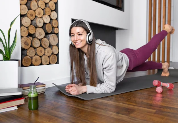 Una-donna sorridente è sdraiata su un-tappetino da-yoga con cuffie, indossa una felpa grigia e leggings viola, con una bevanda verde accanto a-luì.