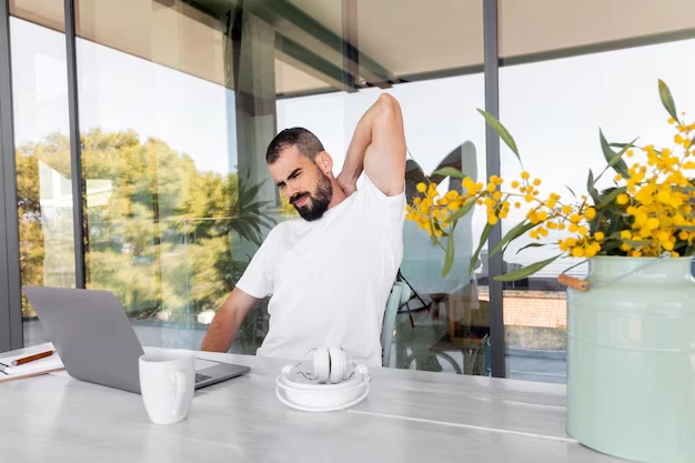 Un-uomo seduto-a un-tavolo allunga-il collo-mentre lavora-su un-portatile, con una-tazza, cuffie, e una-piantina con-fiori-gialli vicina.